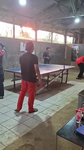 The video shows a table tennis match being played indoors. Three men are visible. In the foreground, a man wearing a black t-shirt and red pants stands with his back to the camera, holding a ping pong paddle. To his right, a man in a blue shirt stands behind the table tennis table. On the other side of the table, a man in a red shirt is in the process of hitting a ping pong ball. The table tennis table is blue with a red trim, and it is set up on a tiled floor. The environment appears to be a covered outdoor area or a rustic indoor space with a wooden ceiling and concrete walls. Natural light streams in from the top of the frame, suggesting it is either morning or late afternoon.