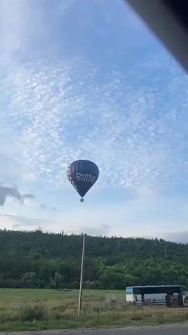 A dark blue hot air balloon with white text that appears to read "Dentus" floats in a partly cloudy sky. The balloon has a checkered pattern on its lower left side. A basket is suspended from the balloon. Below the balloon, a green, tree-covered hill meets a grassy field with a tall wooden pole in the foreground. In the background, a blue and white bus is parked near a fence. The camera is positioned at a low angle, looking up at the balloon, and the bottom edge of the frame shows a dark surface that could be a road or sidewalk. The video consists of a single, static shot.