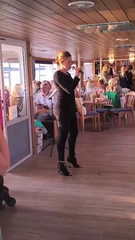 A young woman in a sparkling black jumpsuit and high-heeled shoes sings into a microphone on a wooden floor. She is standing in what appears to be the interior of a boat or a restaurant with a wooden ceiling and walls. In the background, several people are seated at tables, some of whom are looking towards the singer. There is a bar area in the back with bottles on shelves. The lighting is warm and appears to be natural light mixed with indoor lighting.