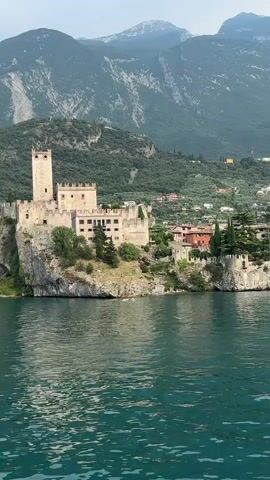 This media file captures a scenic view of a castle perched on a cliff overlooking a large body of water. The castle, made of stone, has a tall tower and a sprawling complex of buildings with a crenelated roofline. Lush green trees grow around the castle, and a smaller, reddish-brown building is visible to its right.

In the background, a steep, verdant mountain rises behind the castle, with more buildings dotting its slopes. The sky is overcast, suggesting a cloudy or misty day. The water in the foreground is a deep, shimmering teal, with ripples indicating a gentle breeze. The overall impression is one of historical grandeur set against a dramatic natural landscape.

The scene appears to be a still shot, possibly taken from a boat, given the perspective of the water. The lighting suggests it is daytime, likely late morning or afternoon, given the diffused light. There is no visible text within the media file. The location is identified as Malcesine, Italy, which is known for its picturesque castle, Castello Scaligero, situated on a promontory on Lake Garda. The image evokes a sense of tranquility and the timeless beauty of the Italian countryside.