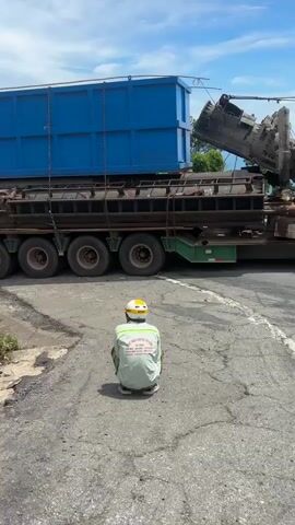 A man in a white shirt and yellow hard hat crouches on a cracked asphalt road. Behind him, a large blue container is loaded onto a multi-wheeled truck trailer. A bundle of large orange hoses is also loaded onto the trailer. To the left of the road, a small pink shrine with a peaked roof sits on a concrete base. The shrine has offerings inside, including bowls and incense sticks. The background shows a hillside covered in grass and trees, with more trucks and cars visible on a road in the distance. The sky is blue with some clouds.