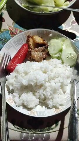 A close-up shot shows a hand forming a heart symbol with its thumb and index finger, positioned in front of a plate of food. The plate contains a portion of white rice, a red sausage, a piece of grilled meat, and several slices of cucumber. Two forks are visible on either side of the plate. The food is arranged on a decorative plate with a geometric pattern around the rim. The plate is set on a table covered with a tablecloth featuring a colorful, patterned design. In the background, another plate with sliced cucumbers can be seen. The lighting suggests an indoor setting, possibly a mealtime. The overall scene captures a moment of presenting or enjoying a meal.