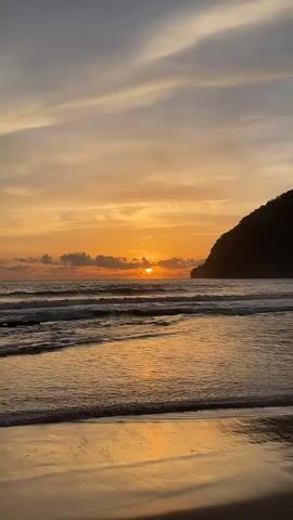 The video shows a sunset over a beach. The sky is orange and yellow, with some clouds. The sun is low on the horizon, and is partially obscured by clouds. The ocean is calm, with small waves breaking on the shore. The beach is sandy, and there is a dark hill or mountain in the background. The video is shot from a low angle, looking out at the ocean. There are no people or objects visible in the video, other than the natural environment.