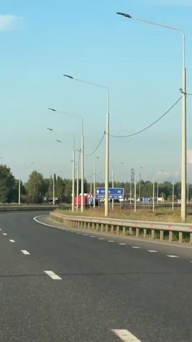 The video shows a view from a moving vehicle on a highway. The road curves to the left, and a metal guardrail runs along the right side. Several tall streetlights with single lamps are visible, spaced along the road. In the distance, trees form a backdrop, and more streetlights and road signs can be seen. A blue road sign points to the right, indicating "PERM" and other destinations with distances. A large truck with a white tank is parked or moving slowly on the left side of the road. The sky is clear blue with a few wispy clouds. The overall environment suggests an open, possibly rural or suburban highway setting.