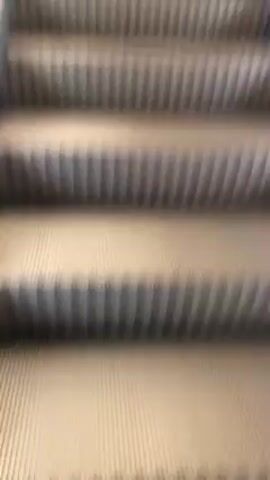 Two men wearing traditional Arab clothing ride an escalator upwards. The escalator has gray steps with vertical ridges, and illuminated white edges. A red sign with white Arabic lettering, and the words "Bank Albilad" in English, is visible on the right side of the escalator. The man closer to the camera wears a gray thobe and a red and white checkered keffiyeh. The man further away wears a white thobe. The escalator is traveling upwards.