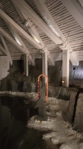 The video shows an underground structure with a body of water and white wooden architecture. The camera slowly pans from right to left. The architecture consists of a low-slung, peaked roof supported by white pillars. Below the roofline on the right side, there is a gray rock formation, upon which there are a few small candles. In front of the formation, a statue stands on a small pedestal, framed by an arched, orange light. The statue and its lighting are reflected in the dark body of water. On the left, there's a white brick wall with an opening leading to a tunnel illuminated by green light. Above the opening, there is a white plaque with text. The reflection of the tunnel and plaque are also visible in the water. The overall lighting is dim, emphasizing the glow of the candles and lights.