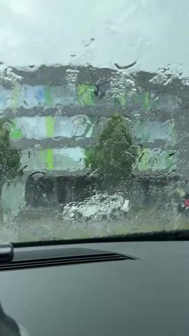 The video shows a view from inside a car in Waldshut, Germany, looking out through a heavily rain-soaked windshield. The car's dashboard is visible at the bottom of the frame, featuring a speaker grille. Outside, the heavy rain creates numerous water droplets and streaks on the glass, significantly blurring the external scene. Through the distorted view, a building with distinct green and blue elements can be seen in the background. Some trees or foliage are also visible, partially obscured by the rain. A white car can be faintly discerned in the distance, further down the road, as the heavy downpour continues. No legible text is visible.