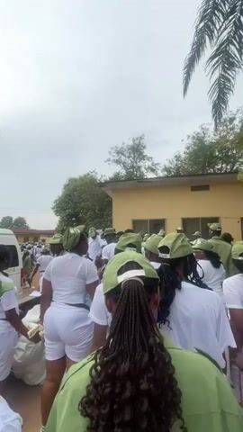 A large group of people, mostly women, are gathered outdoors. They are wearing matching green caps and white outfits. The people are standing in a line, facing a building that has a sign that reads "ORIENTATION CAMP". The sky is cloudy and there are trees in the background. The ground is paved. Some people are carrying bags. The overall impression is of a formal gathering or event.