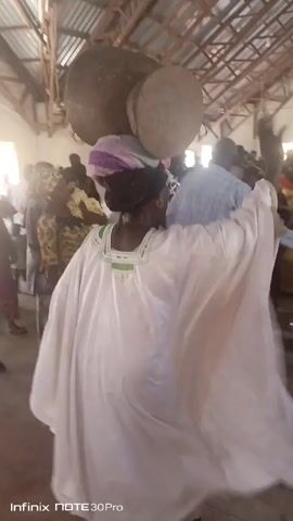 The video shows a woman in a white robe, a purple and white head wrap, and with a large wooden mortar on her head, walking through a crowded room. She holds a bag in her left hand. The room is a large, open space with a metal roof and many people. The camera follows the woman as she walks, capturing her facial expressions, the movement of the mortar, and the other people around her. The lighting is bright and natural. An "Infinix NOTE 30 Pro" watermark is in the bottom left corner.