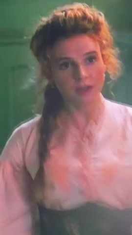 The video shows a woman with red hair styled in braids and curls, wearing a pink Victorian-style dress with lace trim. She is speaking and looking towards the left of the frame. Another woman with long, dark hair and wearing a red skirt and dark top is visible in the background on the right. The environment appears to be indoors, possibly an old building or a set designed to look like one, with a large, dark, industrial-looking door or panel on the right side of the frame. The lighting is dim and green-toned. The red-haired woman extends her left arm, and her hand appears to move in a blurred motion, suggesting she might be touching or interacting with something just out of frame. There is a brief glimpse of what looks like a golden wheel or mechanism on the dark panel. The overall impression is that of a historical drama or period piece.