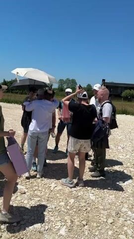 A group of approximately ten people is gathered outdoors on a bright, sunny day with a clear blue sky. They are standing on a light-colored gravel or dirt surface. One person on the far left is partially visible, holding a pink shopping bag. A person in a white long-sleeved shirt and light blue jeans stands with their back to the camera, holding a black bag. Another person in a black t-shirt and patterned shorts is also facing away from the camera, holding a red bag. Several individuals in the group are wearing humorous, brown conical party hats resembling excrement. One person holds a white umbrella over their head for shade. In the background, there are green fields, some sparse trees, and a few distant buildings or structures. The group appears to be engaged in conversation.