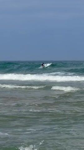 The video shows a surfer riding a wave in the ocean. The water is a mix of green and blue, with white foam from the waves. The sky is a clear blue. There are multiple waves in the ocean, with the surfer on one of the larger waves. The surfer is wearing dark clothing. The video is filmed from a distance, so the surfer's face is not visible.