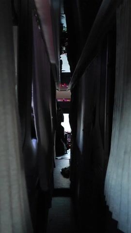 The video provides a low-angle, close-up view looking upward from beneath what appears to be a bed frame or a similar structure. The lighting is dim, with some areas illuminated by faint colored lights, possibly from electronic devices or decor. In the distance, through gaps in the structure, glimpses of objects and possibly people can be seen. One area shows what looks like a stack of books or papers. Another area appears to have some sort of fabric or bedding. The overall impression is of a cluttered, dimly lit space viewed from an unusual, confined perspective.