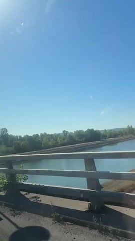 The video shows a view from a bridge over a calm body of water, likely a canal, with lush green trees lining the banks. The sky is a clear, bright blue, suggesting a sunny day. The camera pans slightly, revealing a speed limit sign displaying "40" within a red circle. The scene appears to be in Ottmarsheim, France. The overall mood is peaceful and serene, characteristic of a bright, pleasant day outdoors.