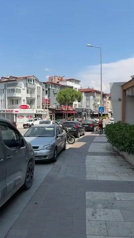 A street scene on a clear, sunny day. The camera is positioned at eye level, looking down a sidewalk next to a street lined with parked cars and buildings. On the sidewalk, a person on a motorcycle is moving away from the camera. The street itself has several cars parked along the side, and a few more are visible further down the road. Buildings on the opposite side of the street appear to be shops or residences, with awnings and balconies visible. The sidewalk is paved with interlocking concrete stones, and a line of green bushes runs alongside the right side of the sidewalk, separating it from a building with a modern facade. The sky is blue with a few wispy clouds.