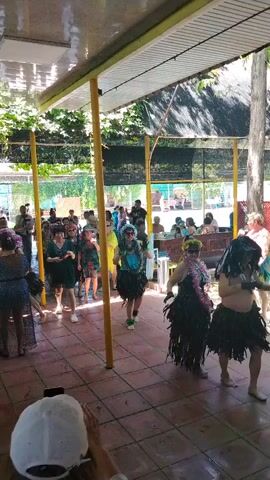 A group of people are gathered outdoors under a covered patio area. In the foreground, a person wearing a white hat is holding up a smartphone to record the scene. To the left, a person in a dark blue dress is visible from the back. Further into the scene, several people are dancing, some wearing costumes that resemble hula skirts and leis. One woman in the center is wearing a yellow outfit and appears to be leading the dance. Other people are standing around watching or participating in the event. The patio floor is tiled. Trees and greenery are visible in the background, suggesting an outdoor setting.