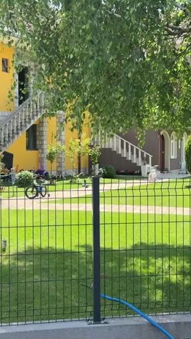 The camera pans from left to right across a lush green lawn behind a black metal fence, focusing on two houses, one yellow and one brown, each with white stairs leading up to a second level.  Various children's toys, including bikes, scooters, and scattered playthings, are visible on the walkway in front of the brown house. The scene is partially shaded by the leafy branches of a tree in the foreground.