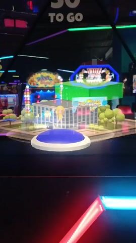 The video shows a brightly lit arcade game with a 3D model of a building surrounded by green trees. The building is light blue with clear glass walls. A blonde, cartoon-like figure is standing on a round, blue platform in front of the building. Above the building are the words "50 TO GO". Other arcade games are visible in the background, with their colorful lights adding to the overall visual spectacle. The video is shot from a low angle, emphasizing the game's features.