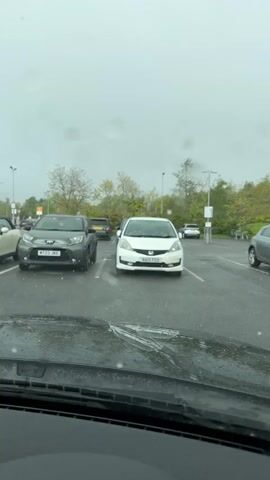 The view from inside a car shows a parking lot on a rainy day. The windshield wipers are in motion, clearing raindrops. Several cars are parked in the lot, including a dark grey car on the left with license plate MT23 JKE, and a white Honda in the center with license plate NA15 FGO. Other cars are visible in the background, parked and likely unoccupied. The sky is overcast and grey. Streetlights and trees are visible in the background.
