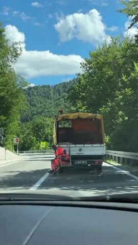 A video taken from inside a vehicle shows the back of a white STRABAG truck with a yellow tarp and orange construction material. A person in an orange high-visibility work suit is sitting on a platform at the back of the truck, holding a white pole with a red cone attached to it. The truck is driving on a paved road with a white line marking the lane. The road is surrounded by lush green trees and a forested mountain in the background. The sky is blue with white clouds. A road sign with a red border and a white background is visible on the left side of the road. Another yellow sign with a black symbol is also visible on the left. The license plate of the truck reads PG FM 136.