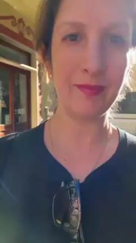 A woman wearing a black long-sleeved shirt and sunglasses is recording a selfie video in a residential area. She has dark hair and is wearing pink lipstick. She is standing on a sidewalk in a neighborhood with buildings of pink and peach tones and small balconies. The buildings line a narrow street that slopes uphill. Several cars are parked along the street. The sky is clear and blue.
