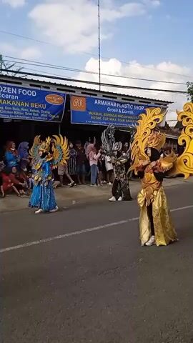 The video shows people in costumes in a parade on a paved street. Three people are in the foreground wearing elaborate outfits with large, decorative, wing-like structures. The costumes are in blue, silver, and gold. The people wearing the costumes are moving forward on the street. A woman in a green shirt, tan skirt, and an orange hat is walking in the opposite direction and holding water bottles. A man in a black vest, a black baseball cap, and sunglasses walks behind the woman. Behind the costumed people, a large, colorful, decorative float on wheels is being pulled along the street. There are other people watching the parade from the side of the road. A blue and white sign hangs on a building in the background advertising a plastic shop. The sky is mostly blue with some white clouds.