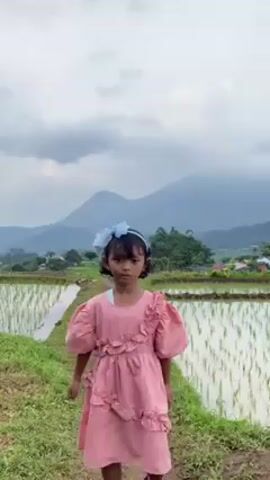 A young girl is standing in a rice field and looks toward the camera, makes peace signs with both hands, and then turns around and runs away. She is wearing a pink dress and a blue bow in her hair. She turns back toward the camera and holds her arms out, then continues to run away. The rice field is surrounded by mountains in the distance. The sky is cloudy.
