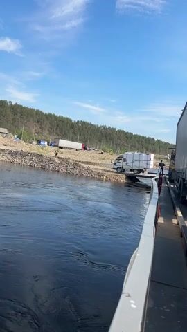 The video captures a sunny day scene with a wide, dark river in the foreground, reflecting the blue sky and white clouds. A ferry, seen from its deck, is partially visible on the right side of the frame, with the railing in focus. On the opposite bank, a gravelly shoreline is lined with trees and a few buildings. Several trucks and vehicles are present, with one white truck loaded with white boxes on a ramp leading into the water. A person is standing near the truck on the ramp. Further down the shoreline, more trucks are parked. The overall impression is one of transportation and activity at a river crossing point.