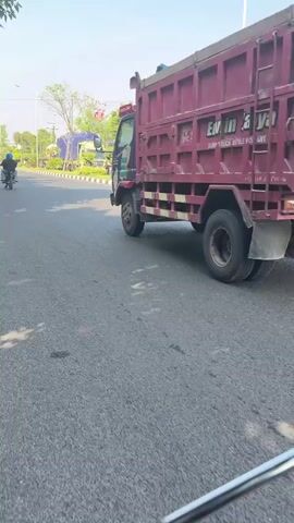 A burgundy dump truck with "EW IN JAYA" written on its side travels on an asphalt road. A person on a motorcycle is visible in the distance to the left of the truck. The environment appears to be a street with some trees and a blue tarp structure in the background. The sun is shining, casting shadows.