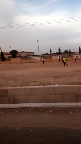 The video captures an outdoor scene, panning horizontally from left to right, revealing a wide, open area that appears to be a sports field or a large dirt ground. The ground is a reddish-brown color, suggesting dry earth or sand. To the immediate right, a paved road is visible, separated from the field by a low concrete curb.

In the background, a large, multi-story building with a stadium-like structure is prominent. It features architectural details such as pillars, arches, and several sections with painted murals or decorative designs. Numerous tall floodlights or streetlights are positioned around the field, indicating its use for various events, potentially at night. A few trees with green foliage are scattered across the field and near the building.

On the field, several individuals are actively engaged in physical exercise. They are wearing brightly colored vests, primarily yellow or neon green, over their clothes. They are seen running or jogging across the dusty surface, suggesting a group training session or athletic activity.

The sky above is bright, with patches of white clouds against a lighter blue or hazy background, indicating daytime. The ambient sound includes significant wind noise, which is consistent with an open, outdoor environment. The continuous camera movement suggests the video was filmed from a moving perspective, possibly from a vehicle or a person walking alongside the field.