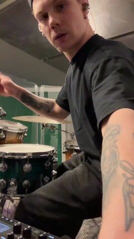 A drummer plays a drum kit in what appears to be a recording studio. The drummer, a young man with a baseball cap, black t-shirt, black pants, and tattoos on his left arm, is seated behind a green and brown drum kit. He is holding drumsticks and hitting the snare drum and cymbal. The studio has soundproofing on the walls and a control board visible to the left of the drummer. Overhead lighting and a clear barrier also appear in the scene. The man is looking towards the camera.