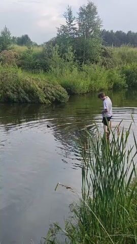 A person wearing a white t-shirt and black shorts stands in shallow water near the bank of a river or lake. They are facing away from the camera and appear to be interacting with something in the water. The surrounding environment is lush and green with trees and tall grass along the bank. The sky is overcast.