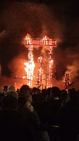 In a dark outdoor setting, flames engulf a lattice-like wooden structure in a bonfire-like event. The fire is intense, with bright orange flames and thick smoke obscuring parts of the structure. A crowd of onlookers, silhouetted against the light of the fire, stands to the right of the frame. Sparks or embers are visible on the ground near the fire. The image is taken from an elevated perspective.