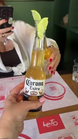 This short video shows two bottles of Corona Extra beer, each with a lime wedge on the neck, being held up, presumably for a toast. The camera focuses on the bottles, with one in the foreground held by a woman's hand with painted nails. In the background, another woman, partially visible, holds a similar bottle. The setting appears to be a restaurant or bar, indicated by the table setting, placemats with "Bento Bar" branding, and a QR code on a coaster. The lighting suggests it is daytime, possibly indoors. The mood appears celebratory or social. Text visible includes "Corona Extra," "La Cerveza," and "Bento Bar." The location context provided is Kryzhanivka, Ukraine.
