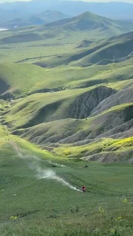 The video shows a scenic landscape featuring rolling, green hills and mountains in the distance. Two people are riding dirt bikes down a grassy hillside, kicking up dust as they move. One rider is wearing a red jacket. The scene gives a sense of adventure and outdoor activity.
