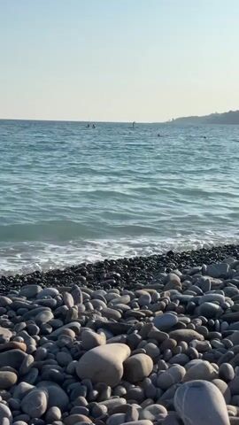 This is a vertical video of a pebble beach on a clear, sunny day. The foreground is dominated by a dense collection of smooth, rounded pebbles in various shades of grey, white, and black. The pebbles are piled up, creating a textured surface that leads down to the water.

The sea is a tranquil turquoise blue, with gentle waves lapping against the shore. The water is slightly choppy, creating a foamy white line where it meets the pebbles. In the distance, several small figures can be seen in the water, appearing to be swimming or enjoying water activities.

Further in the distance, along the horizon, a faint outline of a coastline is visible, suggesting land with some buildings or structures. The sky above is a pale, clear blue, indicating fair weather and likely a warm, sunny day.

The overall mood of the video is peaceful and serene, evoking a sense of a relaxing day at the beach. The focus is on the natural beauty of the coastline, with the gentle movement of the water and the smooth texture of the pebbles. The presence of people in the water suggests a recreational activity, likely swimming or paddleboarding. The time of day appears to be late morning or early afternoon, given the bright sunlight and clear sky. There is no visible text in the video. The location context provided is Ospedaletti, Italy.