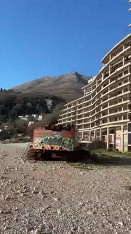 The video shows a large, multi-story, unfinished concrete building. It's a partially built, curved structure with many empty window and balcony spaces. There is graffiti visible on some of the walls and columns. In the foreground, there's a rusted and graffiti-covered construction vehicle, possibly a bulldozer, sitting on a gravelly surface. In the background, there's a mountain range and a clear blue sky. Small buildings can be seen further in the distance near the base of the mountains. The camera tilts upwards, focusing on the unfinished building structure.