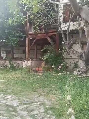 The video shows a traditional Bulgarian house with wooden balconies and stone walls. The house is surrounded by greenery, including trees and bushes. There is a cobblestone path leading to the house. The scene appears to be set in a rural or historic location. The time of day appears to be daytime, with soft lighting suggesting it could be morning or late afternoon. The weather seems pleasant, with no visible rain or harsh sunlight. There are no people visible in the video. The overall atmosphere is peaceful and rustic, evoking a sense of history and tradition. The architecture suggests an older style of construction, typical of historical Bulgarian villages.