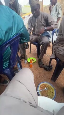 A group of men are sitting on blue plastic chairs, eating food from plates on the ground. The men are dressed in traditional Nigerian clothing, including hats. A plastic bottle of Fanta and a bottle of water sit on the ground. The setting appears to be outdoors, with a sandy ground visible. Some of the men are talking and interacting with each other while they eat. A building is visible in the background.