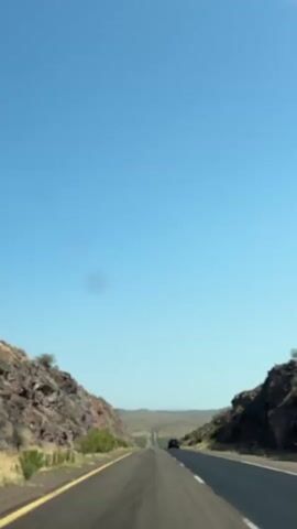 The video shows a view from inside a vehicle driving on a two-lane highway through a desert landscape. The road is asphalt with yellow and white lane markers. Rocky hills covered in sparse vegetation flank the road on both sides. The sky is clear and blue with a few faint clouds. In the distance, the road stretches towards a hazy horizon. Two vehicles are visible on the road ahead, both moving in the same direction as the camera. The first vehicle is a dark SUV or sedan, and the second is a red SUV that is closer to the camera and appears to be overtaking. The red SUV is more prominently featured as it passes. The video captures the sensation of travel and the vastness of the arid environment.