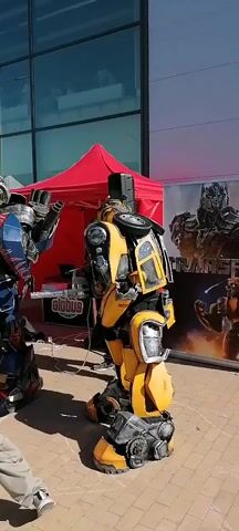 Two individuals dressed in Transformers costumes, one resembling Optimus Prime and the other Bumblebee, are standing outdoors in front of a building with large glass windows. Optimus Prime is on the left, wearing a blue, red, and silver costume with a black helmet. Bumblebee is on the right, in a yellow and black costume, with visible car parts. They appear to be performing or interacting with a small crowd of children who are visible around Optimus Prime. In the background, there is a red tent with a speaker setup and a sign that says "GLOBUS". A large poster featuring Transformers characters is also visible behind Bumblebee. The ground appears to be paved, and the setting suggests a public event or gathering on a sunny day.