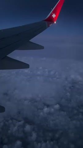 The video presents a view from an airplane window, showcasing the aircraft's wing against a backdrop of dense clouds and a darkening sky. The wing is predominantly grey, and its tip, marked with a red vertical stabilizer, displays a white star emblem. A lit light is also visible on the stabilizer. Below the wing, the dense cloud cover resembles a textured, undulating surface in shades of grey and white. The sky transitions from a dark blue at the top to a lighter greyish hue near the horizon.