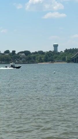 The video shows a boat moving on the water, with a bridge to the right and a water tower in the background. The water is choppy and the sky is partly cloudy. A white buoy floats in the water. Trees line the shore in the background. The boat has a motor and appears to have at least one person in it.