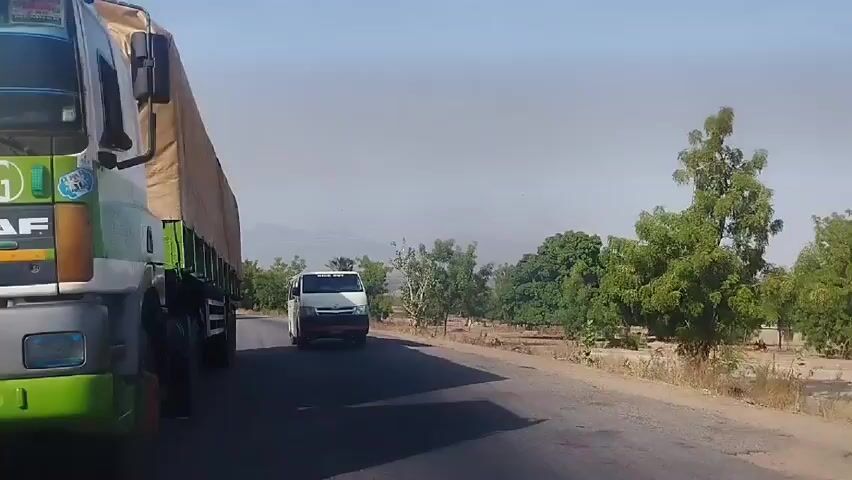 The video shows a road in what appears to be a rural area. The sky is clear and a bit hazy. There are trees and bushes along both sides of the road. In the foreground, a green and white DAF truck with a tan tarp covering its load is moving towards the camera. A white van is also moving in the same direction as the truck but is further down the road. In the distance, a small, red three-wheeled vehicle is also moving in the same direction. A power line tower is visible on the right side of the road.