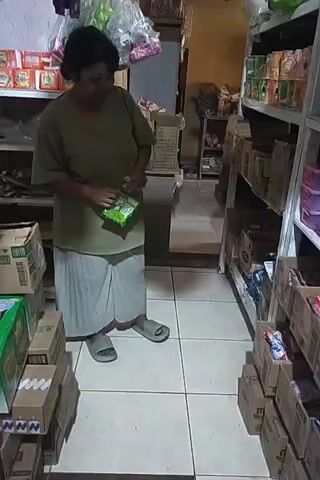 The video shows a person standing in a store aisle filled with shelves of products. The person is wearing a tan t-shirt and white pants. They are holding a small, green package in their hands. To the left of the person, there are stacked boxes, some with green packaging visible. To the right, there are shelves filled with various boxed items. The floor is tiled with a grid pattern. The person appears to be looking down at the package they are holding.