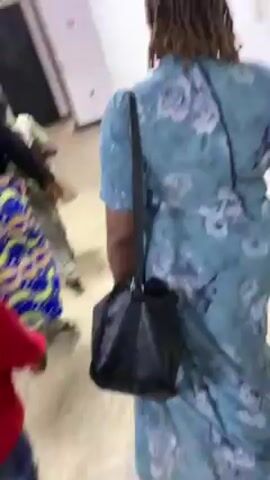 The video shows an indoor space, possibly a polling station or a waiting area, with several people moving around. There are makeshift cubicles with blue curtains visible in the first frame. Later, the camera follows a person walking through a crowded space.  Other individuals are seen moving in different directions, some carrying items such as bags, baskets, or small objects. The lighting is bright, and the general atmosphere appears active.  The colors of clothing worn by the people are varied, including blue, red, yellow, and black. There are white walls and a tiled floor visible.