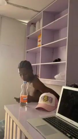 A person standing behind a white counter in a room. The person appears to be shirtless and has their hair in cornrows. On the counter are a bottle of Fanta, a hat, and a laptop. Behind the person is a white shelf unit with items on the shelves. A second shot reveals a person in a gray and white patterned shirt looking at their reflection in a phone. The background features a patterned wallpaper.