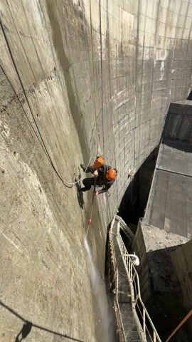 From a high angle, a person is rappelling down the face of a concrete dam, using a pressure washer to clean the surface. The person is wearing a dark suit and an orange helmet, suspended by ropes, with the pressure washer in hand, spraying water down the dam. The dam is curved, with visible vertical striations and wet patches. A metal staircase or walkway is visible along the edge of the dam, reaching into the structure. The sky is visible at the top of the frame, showing a clear, sunny day. Trees are visible at the top of the dam wall.