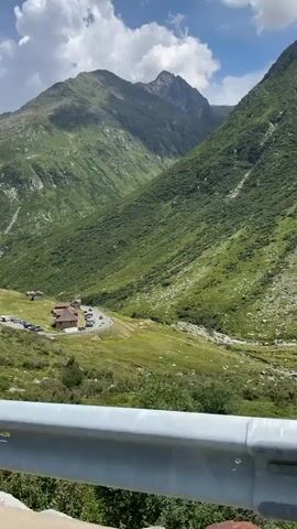 The video shows a scenic view of a lush green mountain valley on a partly cloudy day. In the foreground, a white metal guardrail is visible, mounted on concrete supports. Beyond the guardrail, the landscape slopes down to reveal a winding road with several cars parked near a cluster of buildings. The buildings appear to be a small complex, possibly a restaurant or rest stop, with a brown roof. The valley walls are covered in dense green vegetation, with rocky outcrops and streaks of what could be water runoff visible on the steeper slopes. The mountains in the background are large and imposing, with a peak reaching towards the sky. The overall impression is one of natural beauty and remoteness.