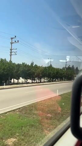 The video shows a view from a vehicle moving along a road. The road is paved and has a white line marking the lanes. On the left side of the road, there is a line of trees. Beyond the trees, there are power lines and some industrial structures. On the right side of the road, there is grassy terrain with some bushes and trees. The sky is clear and blue, indicating a sunny day. Various vehicles, including cars and trucks, are visible on the road and parked on the side. A Turkish flag is also seen on a pole. The overall scene suggests a journey through a semi-urban or industrial area on a bright, clear day.